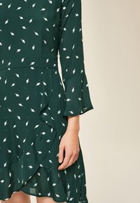 Vestido verde con patrón de hojas blancas, mangas acampanadas y dobladillo con volantes. Hecho de una tela ligera, con cintura ajustada y silueta fluida.