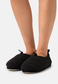 Black slip-on slippers made of soft, textured fabric. Features a round toe, elastic accents, and a contrasting black sole.