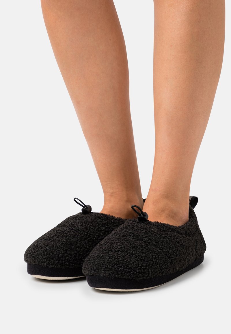 Black slip-on slippers made of soft, textured fabric. Features a round toe, elastic accents, and a contrasting black sole.