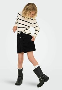 Bambina che indossa una gonna nera di velluto a coste, un maglione a righe bianche e nere, calze bianche e stivali neri, in posa con una mano sui fianchi e che guarda in basso.