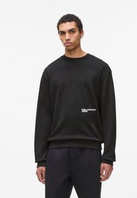 Vīrietis ar īsiem, viļņainiem matiem, ģērbies melnā kapucē ar uzrakstu "Karl Lagerfeld Jeans" priekšpusē, un melnās biksēs uz baltas fona.