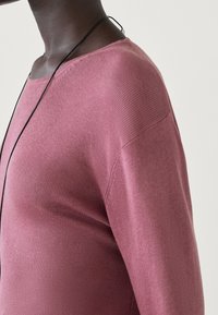 Langslavige, geribbelde trui in gedempt roze met een ronde halslijn. Heeft een comfortabele pasvorm en een soepele textuur. Dunne zwarte koordketting zichtbaar.