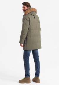 Langer grüner Pufferjacke mit einem mit Fell gefütterten Kapuze, Reißverschluss vorne, seitlichen Schlitzen und Logo-Patch. Getragen über blauen Jeans und braunen Stiefeln.