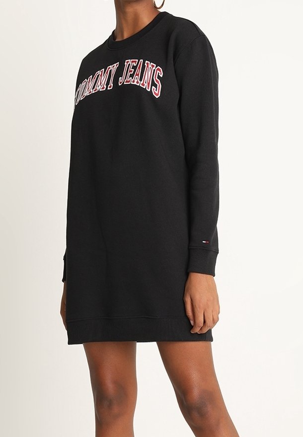 Black sweatshirt dress with "TOMMY JEANS" logo in red and white across the chest. Features long sleeves and ribbed cuffs. Simple design.