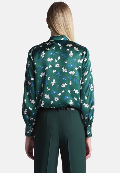 Femme aux cheveux blonds et raides portant un chemisier vert à manches longues avec un imprimé floral bleu et blanc et un pantalon vert foncé, vue de dos.