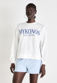 Bela majica s kapuco z modrim napisom "MYKONOS BLUE OCEAN", okrogli ovratnik, rebraste manšete, oblečena z svetlomodrimi kratkimi hlačami. Gladka tekstura tkanine.