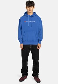 Giovane in felpa blu con il testo "VENTO ACQUA FUOCO TERRA", jeans neri e sneakers rosso scuro, in piedi su uno sfondo semplice.