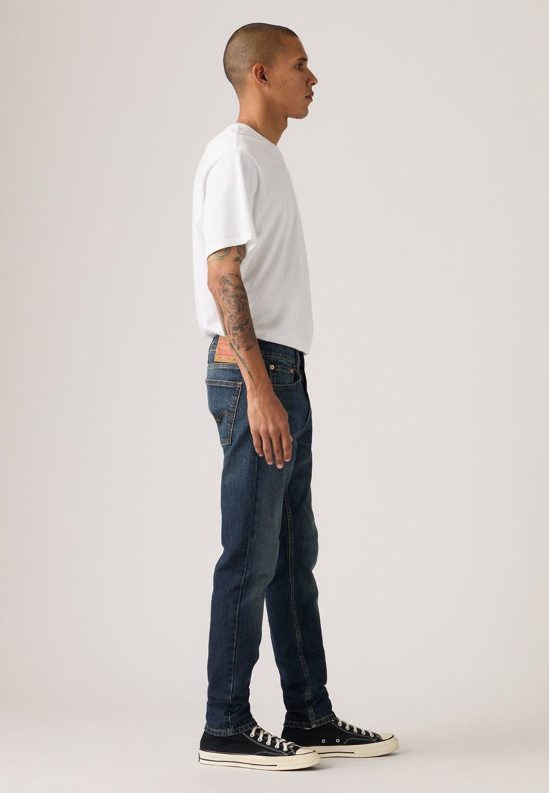 T-shirt blanc à manches courtes ; jean bleu ; baskets noires avec semelles blanches ; tatouage visible sur le bras ; vue de profil debout.