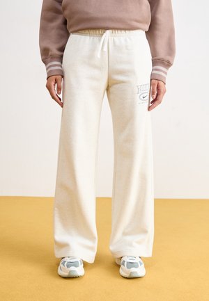 Osoba ubrana w kremowe szerokie dresy z logo Nike, beżową bluzę z paskowanymi mankietami i białoszare buty sportowe stojąca na musztardowej podłodze.