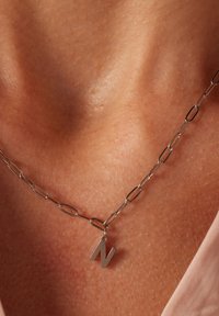 Zilveren ketting met verlaagde schakels en een minimalistische 'N' charm als het belangrijkste aandachtspunt, gepresenteerd tegen een huidskleurige achtergrond.