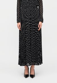 Μαύρη πλισέ φούστα με λευκές πουά βούλες, κατασκευασμένη από ελαφρύ ύφασμα, συνδυασμένη με μαύρα μυτερά τακούνια. Ορατό μακρυμάνικο πουά τοπ.