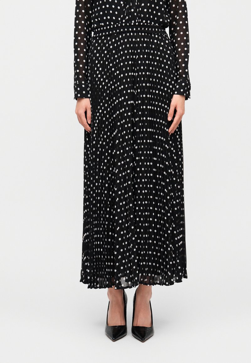 Μαύρη πλισέ φούστα με λευκές πουά βούλες, κατασκευασμένη από ελαφρύ ύφασμα, συνδυασμένη με μαύρα μυτερά τακούνια. Ορατό μακρυμάνικο πουά τοπ.