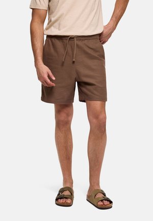Hombre con pantalones cortos marrones con cordón y sandalias oliva con hebilla, de pie contra un fondo claro.