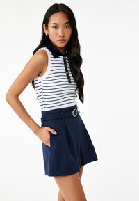 Ærmeløs polo top med hvide og marineblå striber, ribbet tekstur og en marineblå krave. Kombinationen med højtaljede marineblå shorts med bæltestropper.