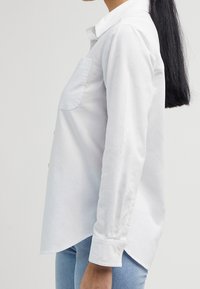 Chemise blanche boutonnée en coton avec manches longues, une poche sur le côté gauche de la poitrine et un ourlet arrondi. Dotée de fermetures à boutons aux poignets.