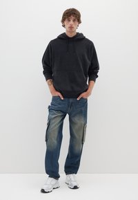 Felpa nera con tasca frontale, vestibilità ampia, abbinata a jeans cargo azzurri chiari con tasche laterali e un finish sbiadito. Sneakers bianche completano il look.