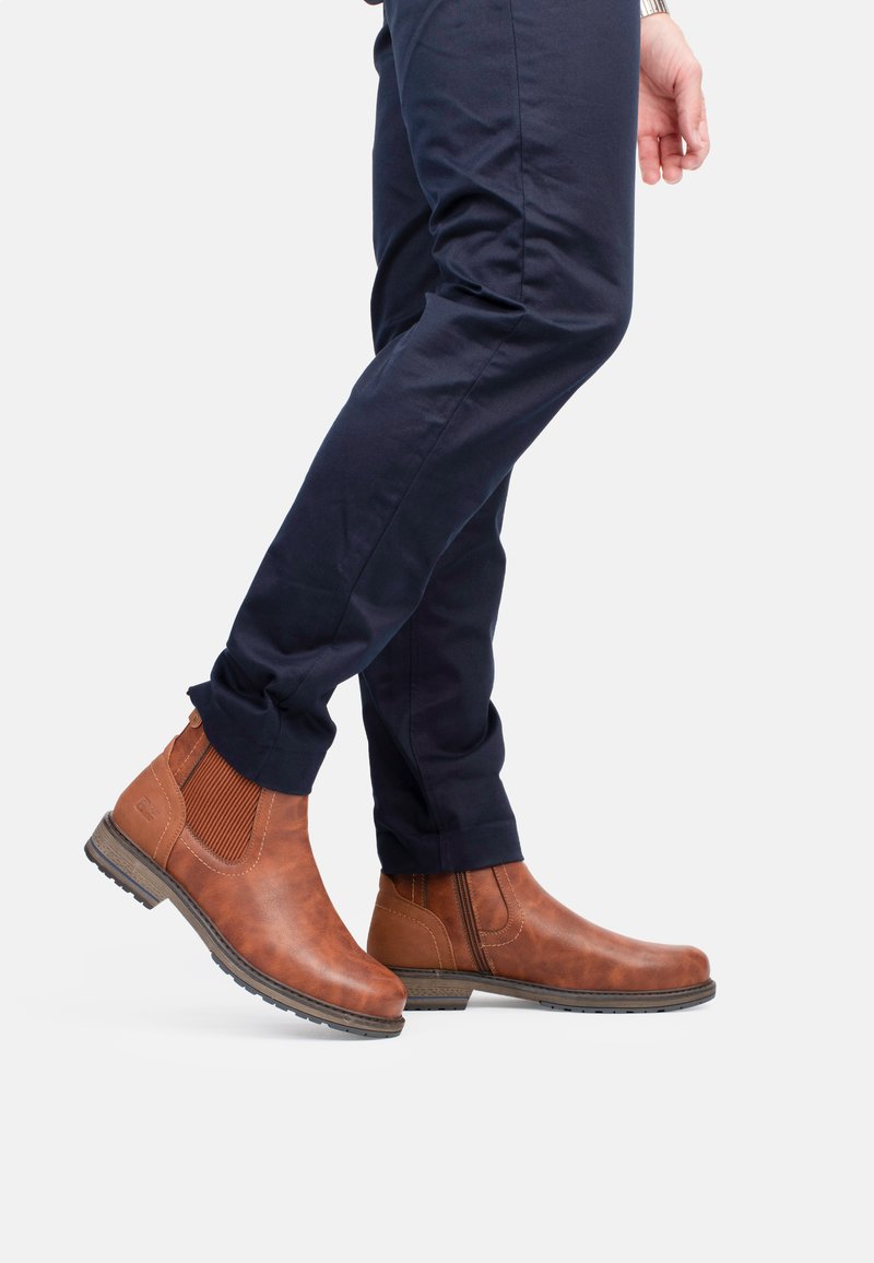 Bottines Chelsea en cuir marron avec des panneaux latéraux élastiques et des accents texturés. Semelle en caoutchouc robuste. Portées avec un pantalon bleu marine.