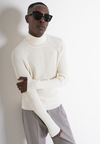 Maglione a collo alto a coste crema con maniche lunghe, abbinato a pantaloni grigi. Modella con occhiali da sole neri angolari. Sfondo semplice.