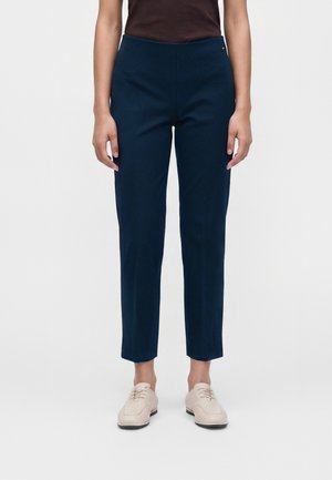 Personne portant un pantalon slim bleu foncé et des chaussures à lacets beige clair, debout devant un fond uni.