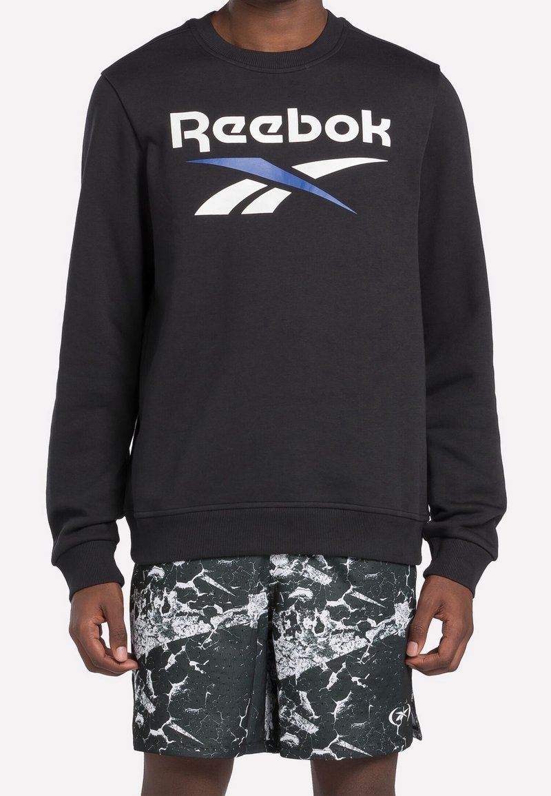 Personne portant un sweat-shirt Reebok noir avec un logo blanc et bleu et un short de sport noir et blanc marbré.