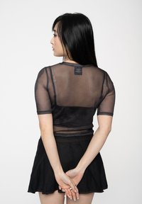 Young woman with long black hair wearing a black sheer mesh top over a black bralette and a black skirt, standing with hands clasped behind back.