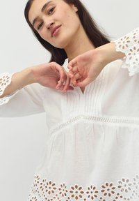 Blusa bianca con maniche lunghe caratterizzata da motivi floreali ricamati lungo l'orlo e dettagli in pizzo. Tessuto leggero con pieghe verticali sul collo.