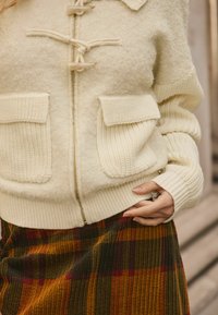 Cream sweater with zip closure, textured fabric, ribbed cuffs, and two front pockets. Paired with a pleated, plaid skirt in autumn colors.