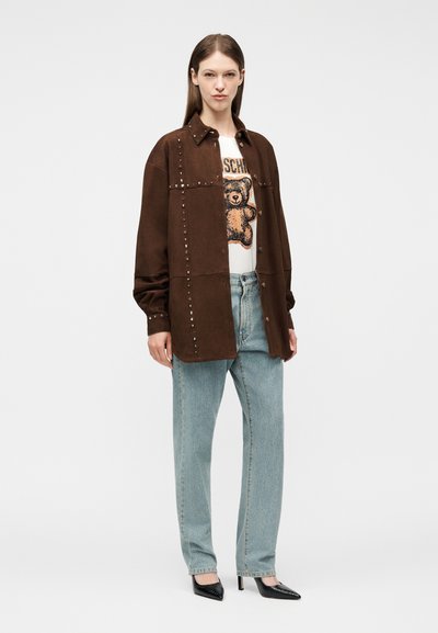 Γυναίκα που φοράει καφέ μπουφάν με τρουκς πάνω από λευκό t-shirt με τύπωμα αρκουδάκι, ανοιχτόχρωμο μπλε τζιν και μαύρες μυτερές γόβες, όρθια σε λευκό φόντο.