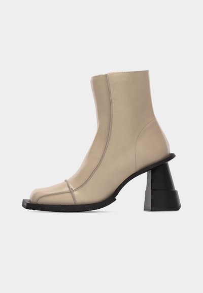 Bottine beige en cuir avec bout carré, couture visible, et talon bloc noir sculpté et épais sur fond blanc.