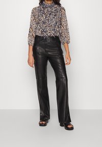 Pantaloni neri in pelle a gamba dritta abbinati a una blusa stampata con collo alto e maniche a sbuffo in tonalità calde di blu, arancione e crema.