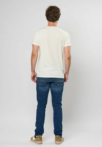 Krátký rukáv krémového trička, spárovaný s modrými džíny. Neformální obuv doplňuje outfit, s hnědou barvou a černými akcenty.
