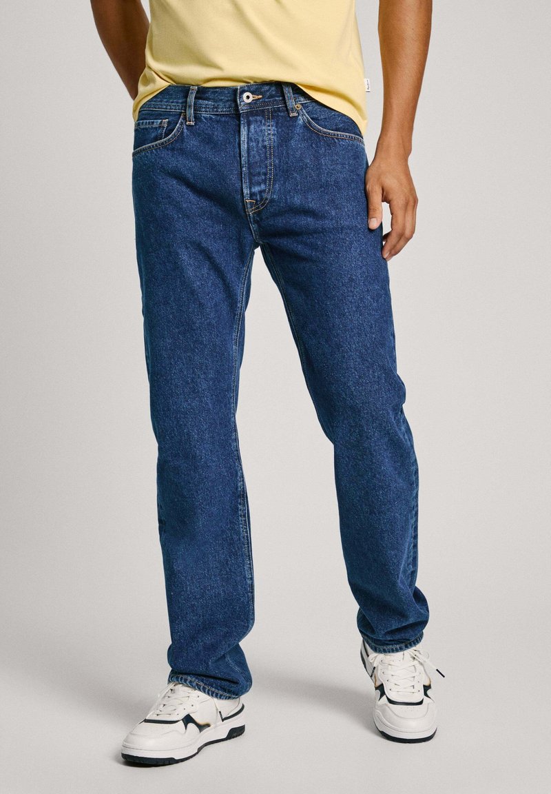 Jeans de denim azul con un diseño clásico de pierna recta, cinco bolsillos, costuras en contraste y un aspecto desgastado, combinados con zapatillas blancas.