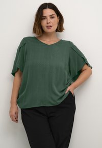 Grøn kortærmet top med V-hals, der har et tekstureret diamantmønster. Toppen er kombineret med sorte bukser, har en afslappet pasform og brede ærmer.