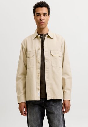 Homme aux cheveux courts et bouclés, portant une veste beige boutonnée sur une chemise foncée et un jean noir délavé, debout devant un fond clair uni.