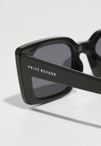 Čierne obdĺžnikové slnečné okuliare s tmavými šošovkami. Rám z acetátu má ostré hrany a strieborné logo "PRIVÉ REVAUX" na ramene.