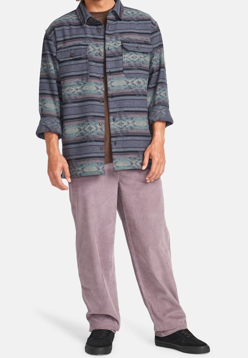 Homme portant une chemise rayée boutonnée avec les manches retroussées, pantalon en velours côtelé mauve, baskets noires, debout sur un fond neutre.