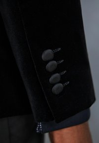 Manga de un blázar de terciopelo negro con cuatro botones texturizados, cada uno con un acabado suave. Detalles de costura a lo largo del borde de la manga.