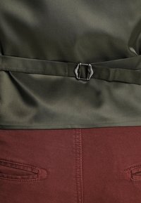 Cintura in tessuto verde oliva con accessori in argento, fissata attorno a un indumento color bordeaux, con tasche dettagliate da cucitura e una texture liscia.