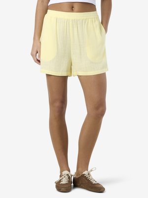 Nő világos sárga elasztikus derekú rövidnadrágot visel elülső zsebekkel és barna fűzős sportcipővel, fehér háttér előtt áll.