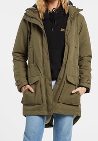 Parka vert olive avec fermeture à glissière et à boutons-pression, poches avant et capuche, portée sur un sweat à capuche noir et un jean bleu clair.