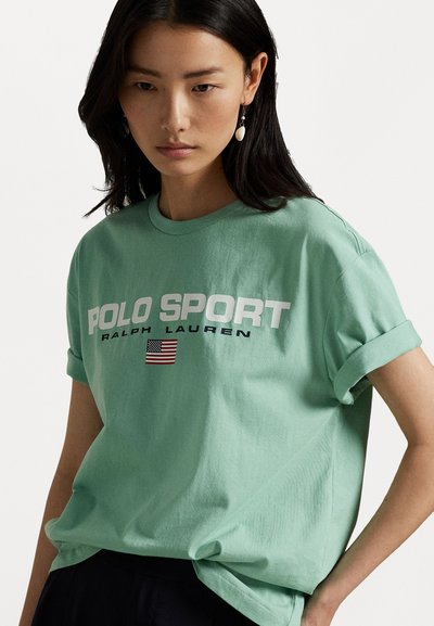 Zaļš kokvilnas T-krekls ar sarullētām piedurknēm, uz kura redzams balts "POLO SPORT RALPH LAUREN" teksts un neliela Amerikas karoga grafika.