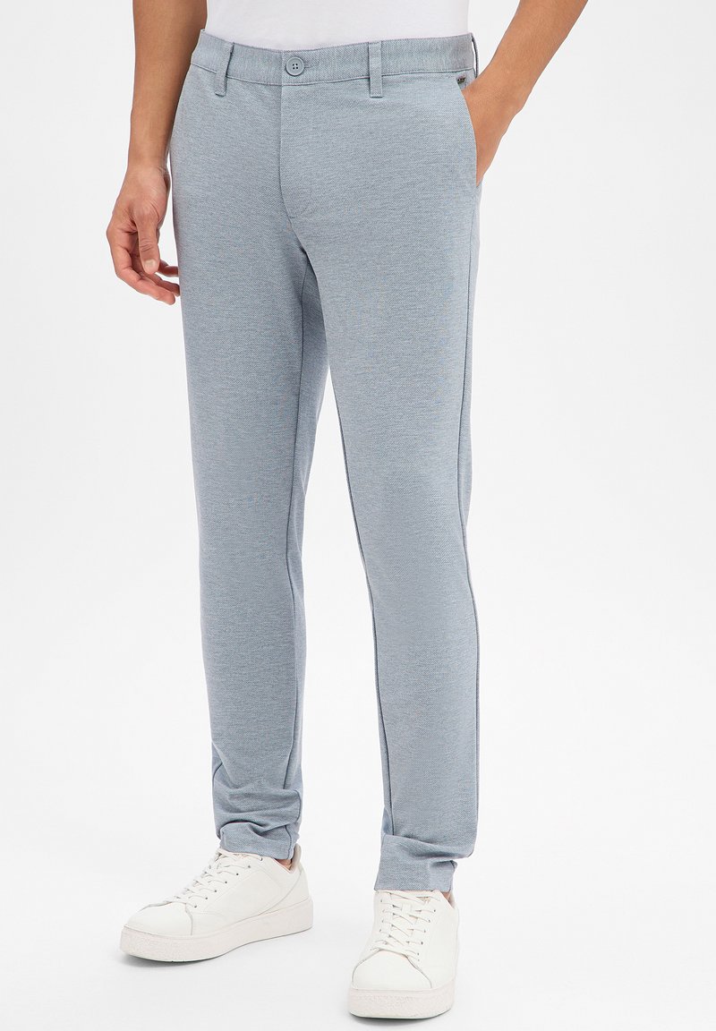 Pantalon de survêtement bleu clair ajusté avec un tissu texturé et une fermeture à bouton. Porté avec des baskets blanches, une main dans la poche.