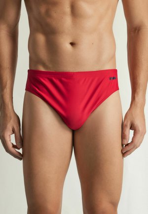 Torace maschile con slip da bagno rosso tinta unita, in piedi davanti a uno sfondo chiaro semplice, mani rilassate ai lati.