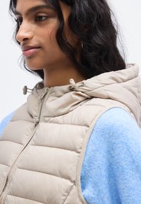 Μπεζ αμάνικο μπουφάν με κουκούλα, διαθέτει εμπρόσθιο φερμουάρ και ραμμένα πάνελ. Φορά ται πάνω από ένα ανοιχτό μπλε πουλόβερ με μαλακή υφή.