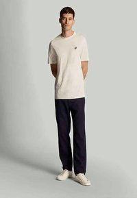Λευκό βαμβακερό t-shirt με κοντά μανίκια, με μικρό λογότυπο στο στήθος, σε συνδυασμό με ναυτικές παντελόνες και λευκά αθλητικά παπούτσια.