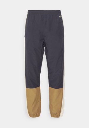 Tmavomodré tréningové nohavice s elastickým pásom, béžovými bočnými pruhmi a hnedou dolnou časťou; vyrobené z ľahkej, hladkej látky.