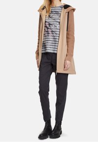 Long beige coat with ribbed sleeves, grey striped shirt with graphic detail, black pants, and black ankle boots. Smooth and textured materials.