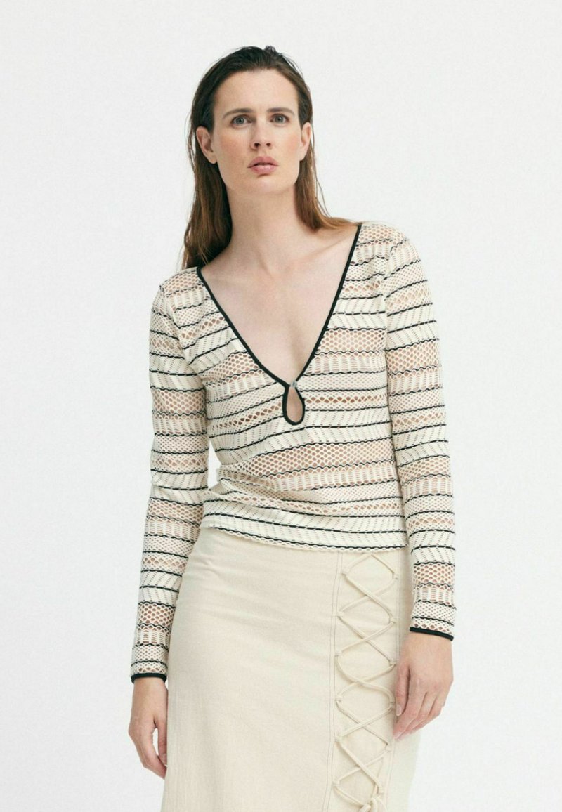 Langärmeliges Oberteil in Creme mit schwarzen und pfirsichfarbenen horizontalen Streifen, tiefem V-Ausschnitt und einem Schlüsselloch-Detail. Kombiniert mit einem beigen Rock mit Schnürung.