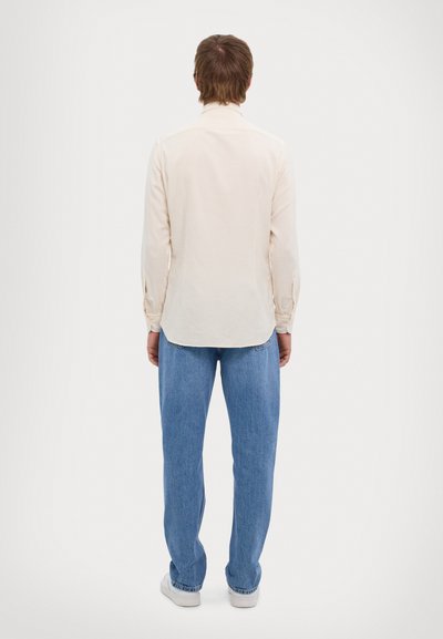 Camisa de manga larga color beige claro con cuello, puños abotonados y dobladillo curvado, combinada con jeans anchos azules y zapatillas blancas.