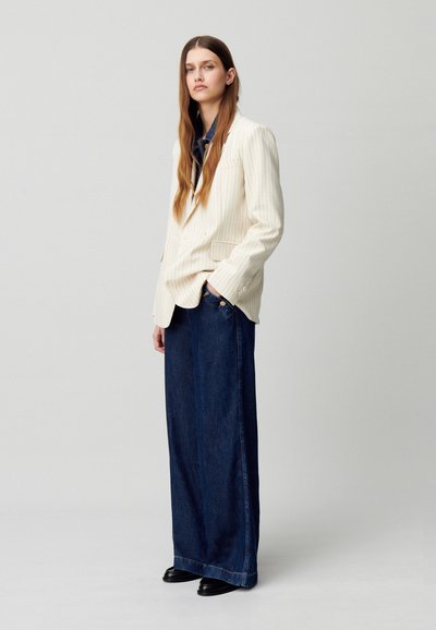 Blazer a righe crema con due bottoni indossato sopra una maglietta blu navy, abbinato a pantaloni di jeans scuri a gamba larga e scarpe nere. Design semplice, vestibilità su misura.